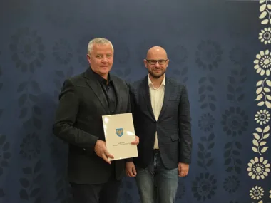 Dwaj mężczyźni stoją obok siebie przed granatową ścianą w kwiaty, jeden z nich trzyma dokument z herbem.