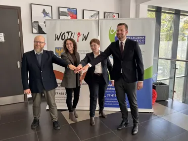 Cztery osoby w eleganckim stroju stoją w holu przed banerami i łączą dłonie w geście współpracy.