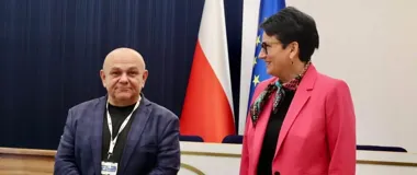 Mężczyzna w niebieskiej marynarce i kobieta w różowej marynarce stoją obok siebie przed flagami Polski i Unii Europejskiej w sali konferencyjnej.