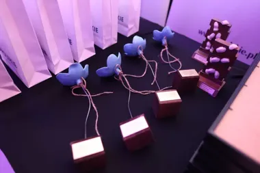 Na czarnym stole pod fioletowym światłem ustawiono białe torby, niebieskie figurki w kształcie kwiatów na drewnianych podstawkach oraz brązowe statuetki z rzeźb